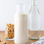 A prepared jar of cashew milk, with a carafe of filtered water and a bowl of raw cashews in the background.