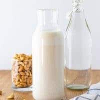 A prepared jar of cashew milk, with a carafe of filtered water and a bowl of raw cashews in the background