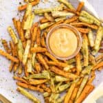 The ultimate crispy baked sweet potato fries on a baking sheet surrounding a bowl of chipotle aioli.