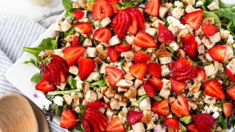 A large square white platter of strawberry goat cheese salad with the dressing on the side and wooden serving spoons.