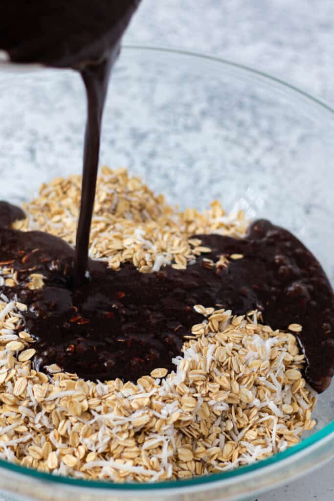Pouring the cocoa butter sauce overtop of the oats and shredded coconut in a large mixing bowl.