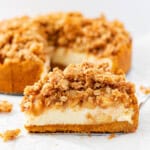 A single piece of apple crumble cheesecake on a silver cake server with the rest of the cheesecake in the background.