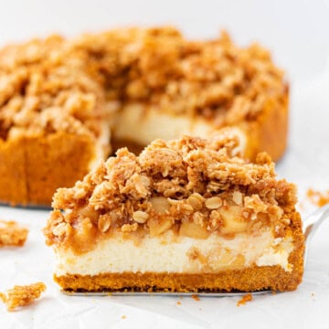 A single piece of apple crumble cheesecake on a silver cake server with the rest of the cheesecake in the background.