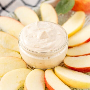 A jar of apple dip in the center of a plate with apple slices surrounding it.