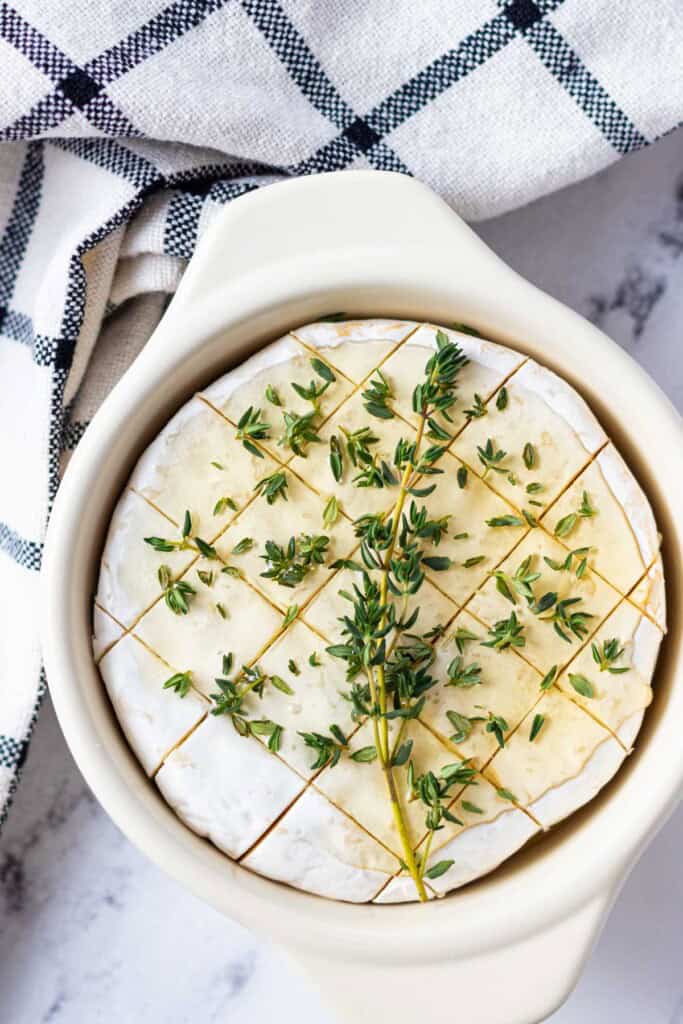 Scored camembert cheese topped with honey and fresh thyme in a brie baker.