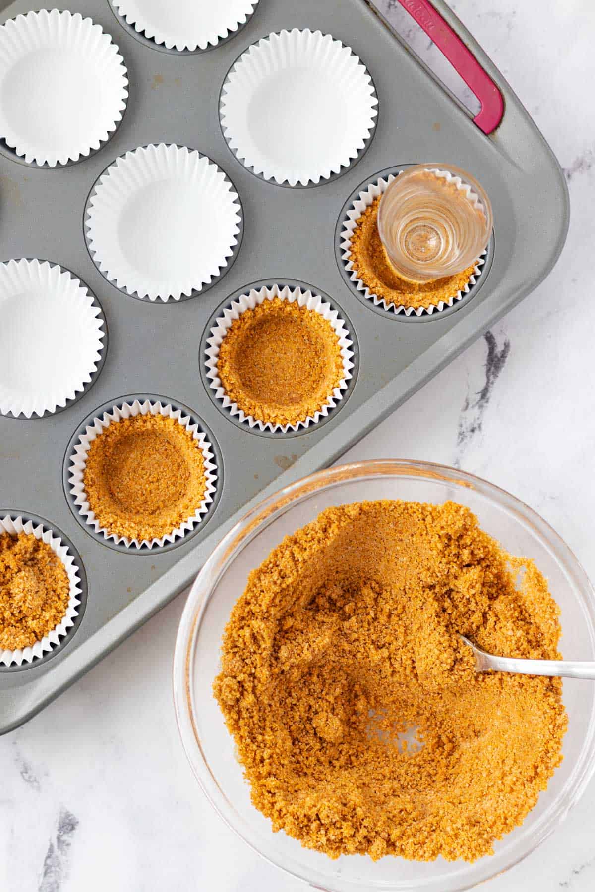 Filling each cupcake liner with graham cracker crust and pressing it down with a shot glass.