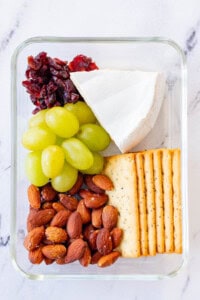 Brie, cranberry, grape, and roasted almond lunchable in a glass container.