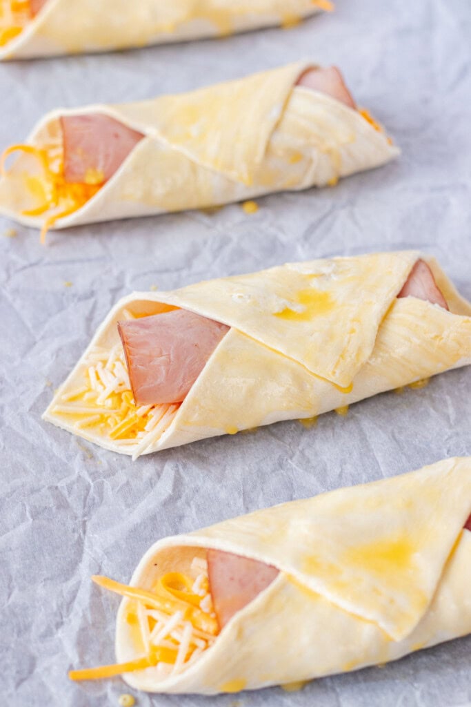 Croissants wrapped with ham, cheese and dijon inside on baking sheet with parchment paper and brushed with egg.