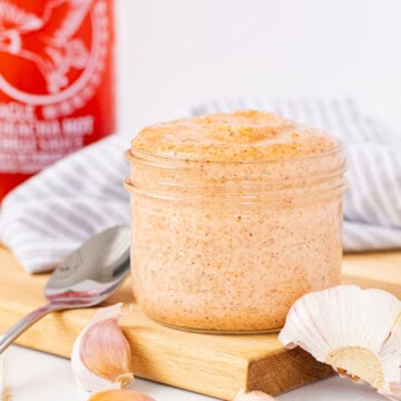 A jar of sriracha aioli on a wooden cutting board with garlic cloves and a bottle of sriracha hot sauce in the back.