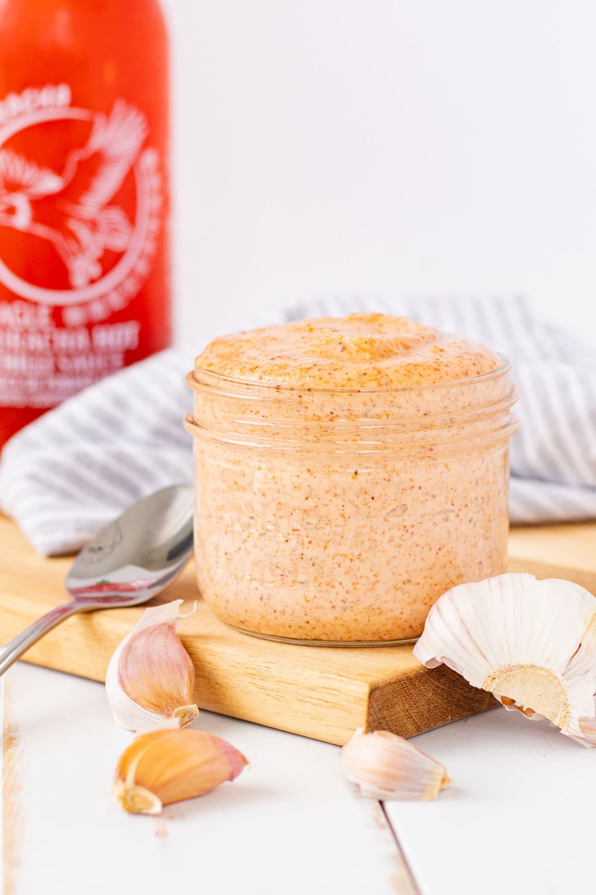 A jar of sriracha aioli on a wooden cutting board with garlic cloves and a bottle of sriracha hot sauce in the back.
