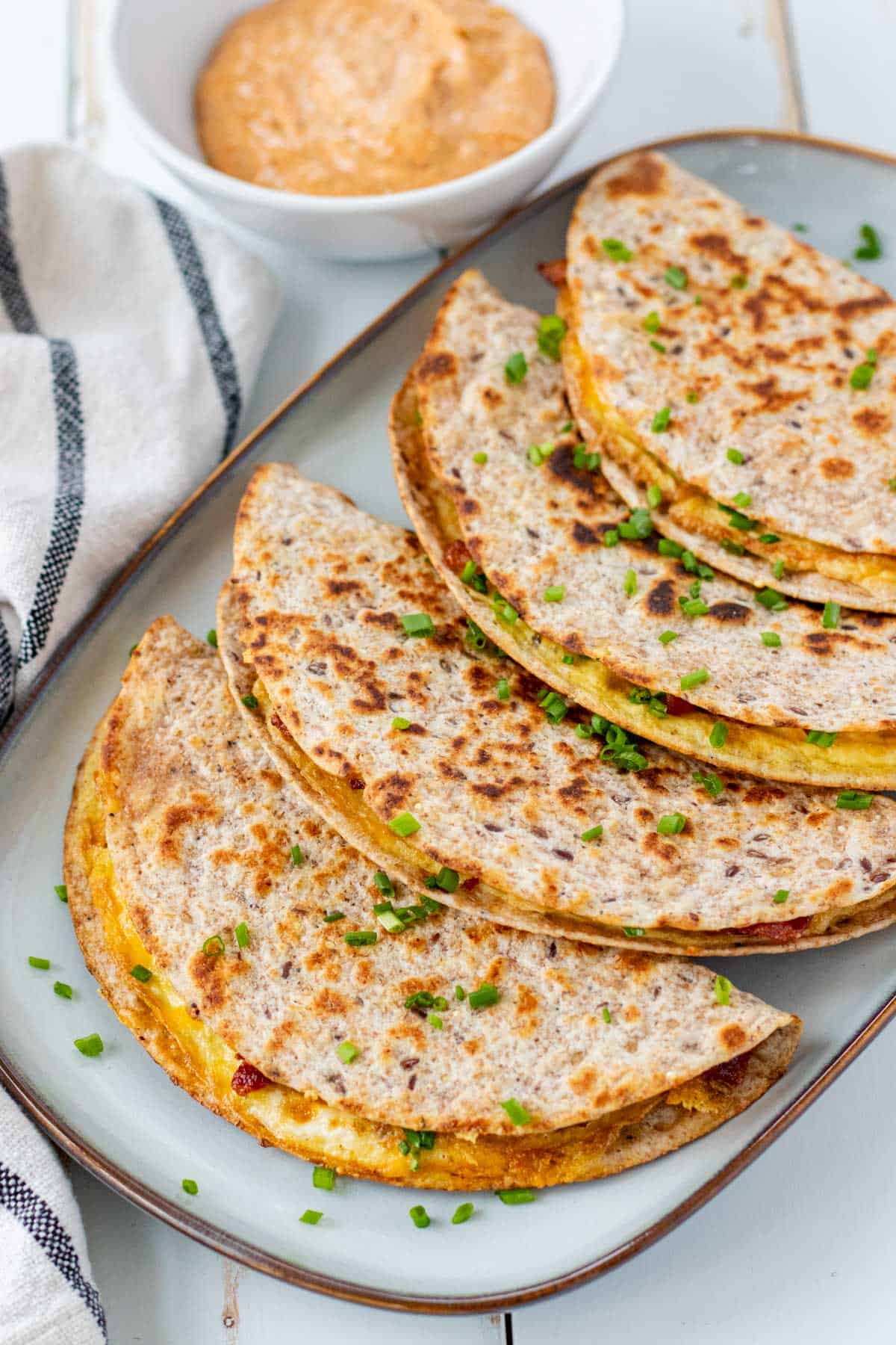 Breakfast tacos lined up on a blue serving plate with a bowl of sriracha aioli in the background.