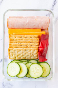 Ham and cheese lunchable with cucumber slices in a glass container.