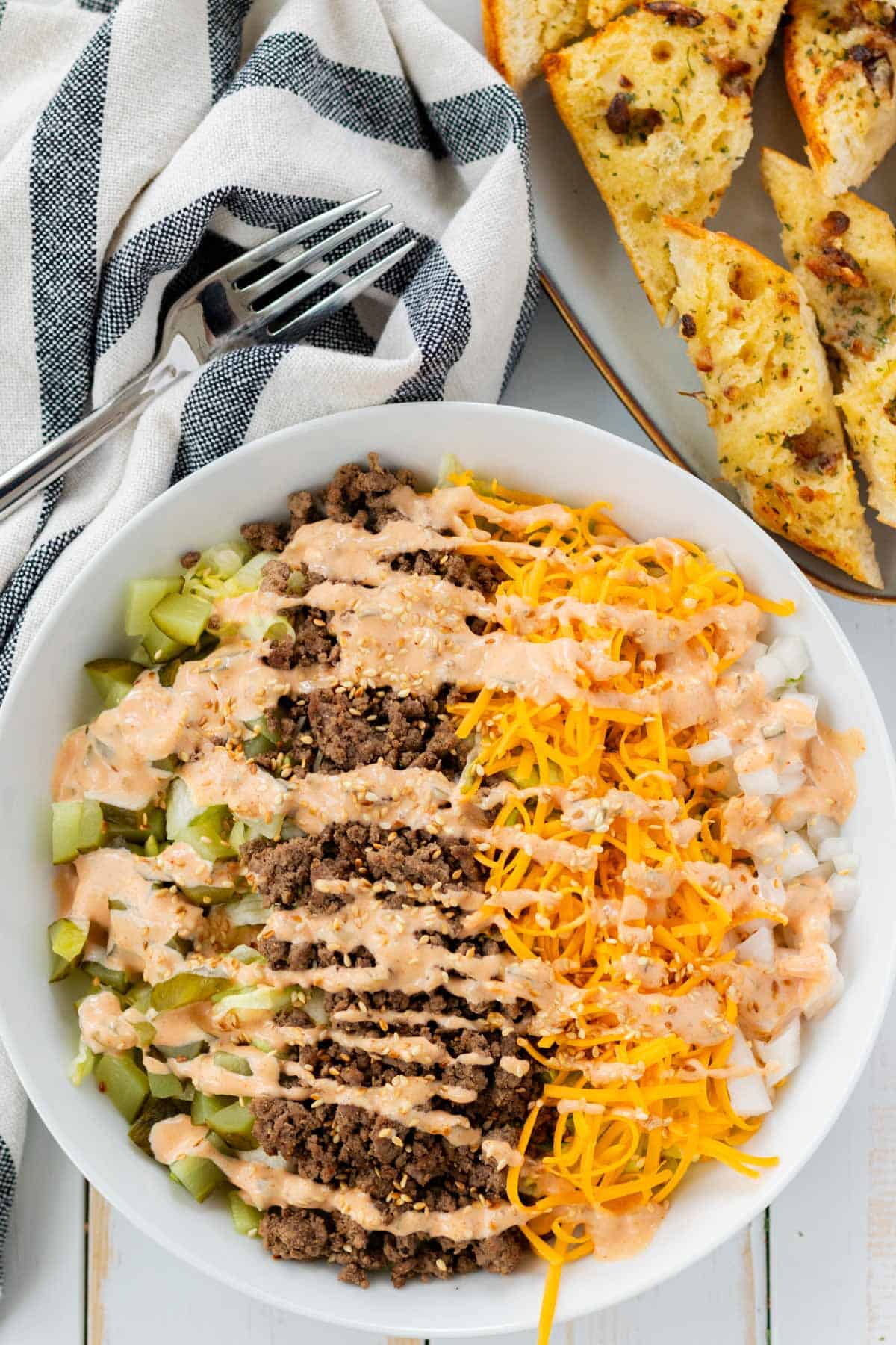 A large big mac salad in a white bowl with garlic bread baguette on the side.