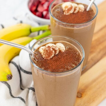 Chocolate peanut butter banana smoothies in two glasses, sprinkled with cocoa powder and topped with extra sliced bananas.