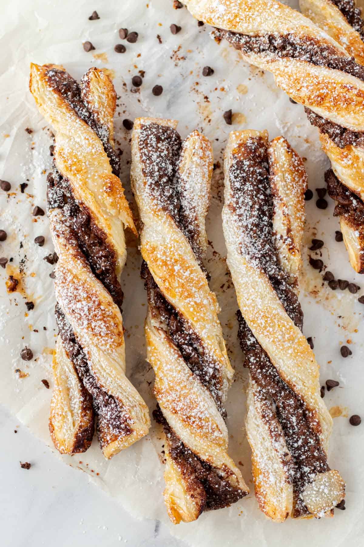 Three individual chocolate puff pastries on a piece of parchment paper with mini chocolate chips sprinkled in the background.