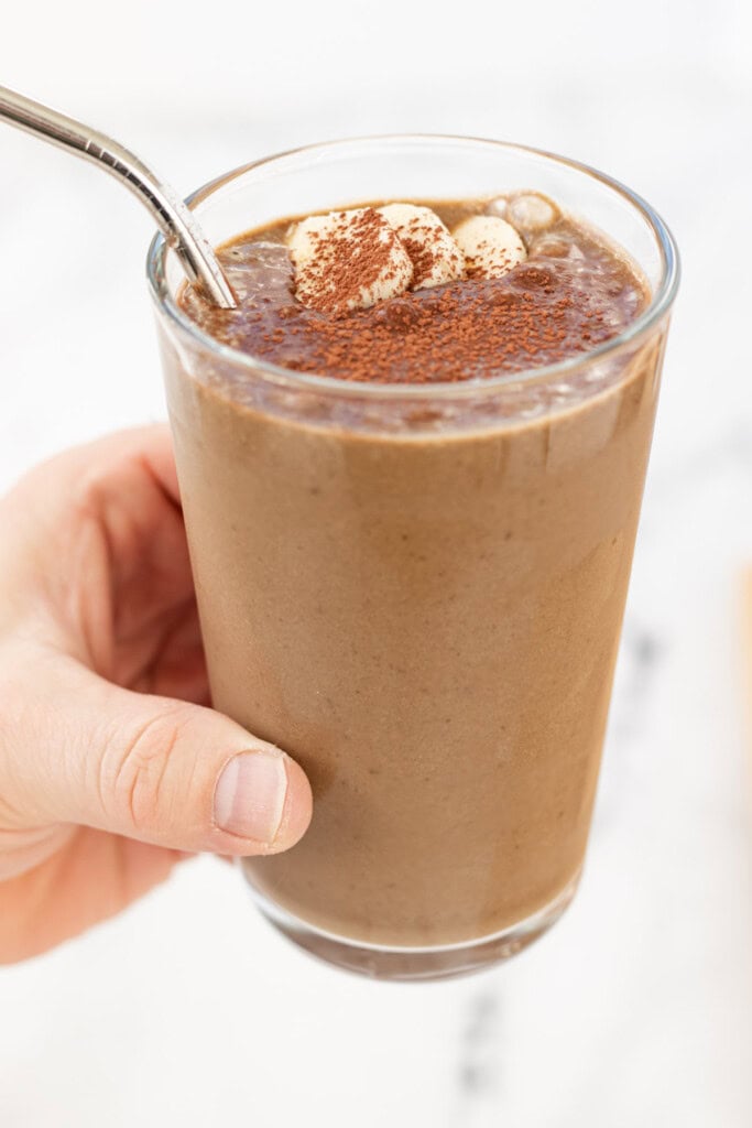 A hand holding a glass of banana chocolate smoothie with a steel straw.