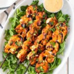 Grilled bang bang shrimp skewers on a white plate set on top of a bed of arugula with a pot of bang bang sauce on the side.