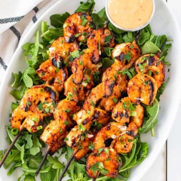 Grilled bang bang shrimp skewers on a white plate set on top of a bed of arugula with a pot of bang bang sauce on the side.