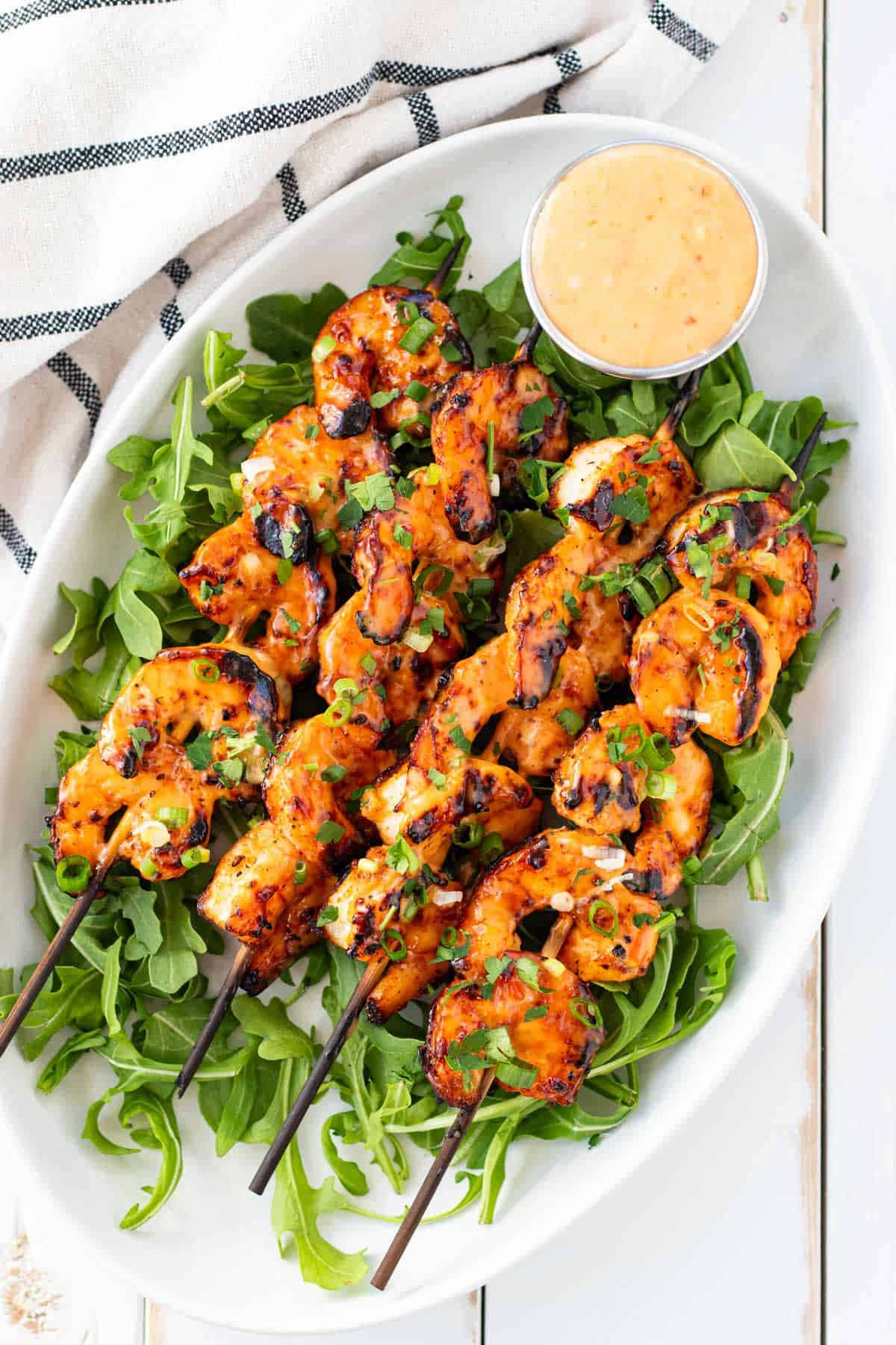 Grilled bang bang shrimp skewers on a white plate set on top of a bed of arugula with a pot of bang bang sauce on the side.
