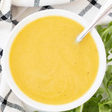 A bowl full of honey dijon salad dressing and a spoon with arugula leaves on the side.