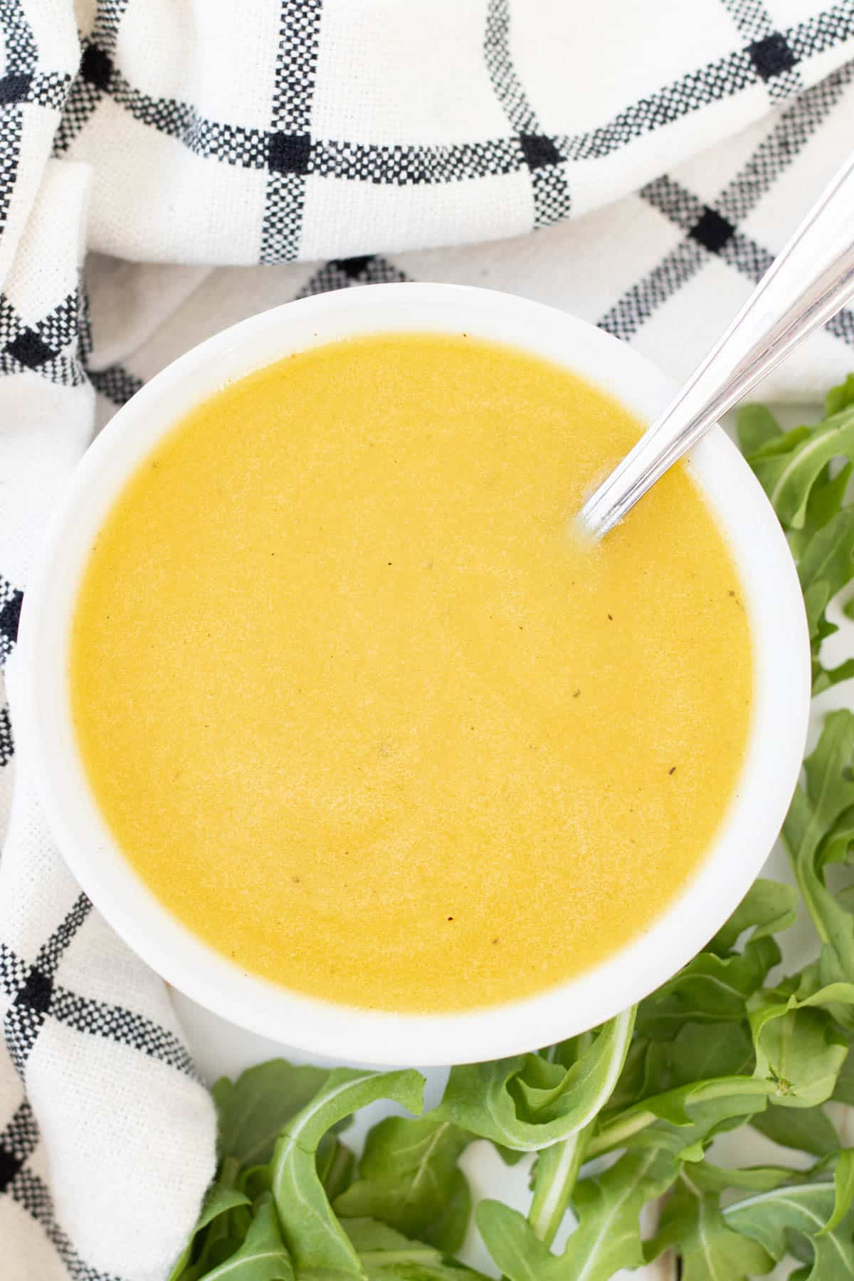 A bowl full of honey dijon salad dressing and a spoon with arugula leaves on the side.