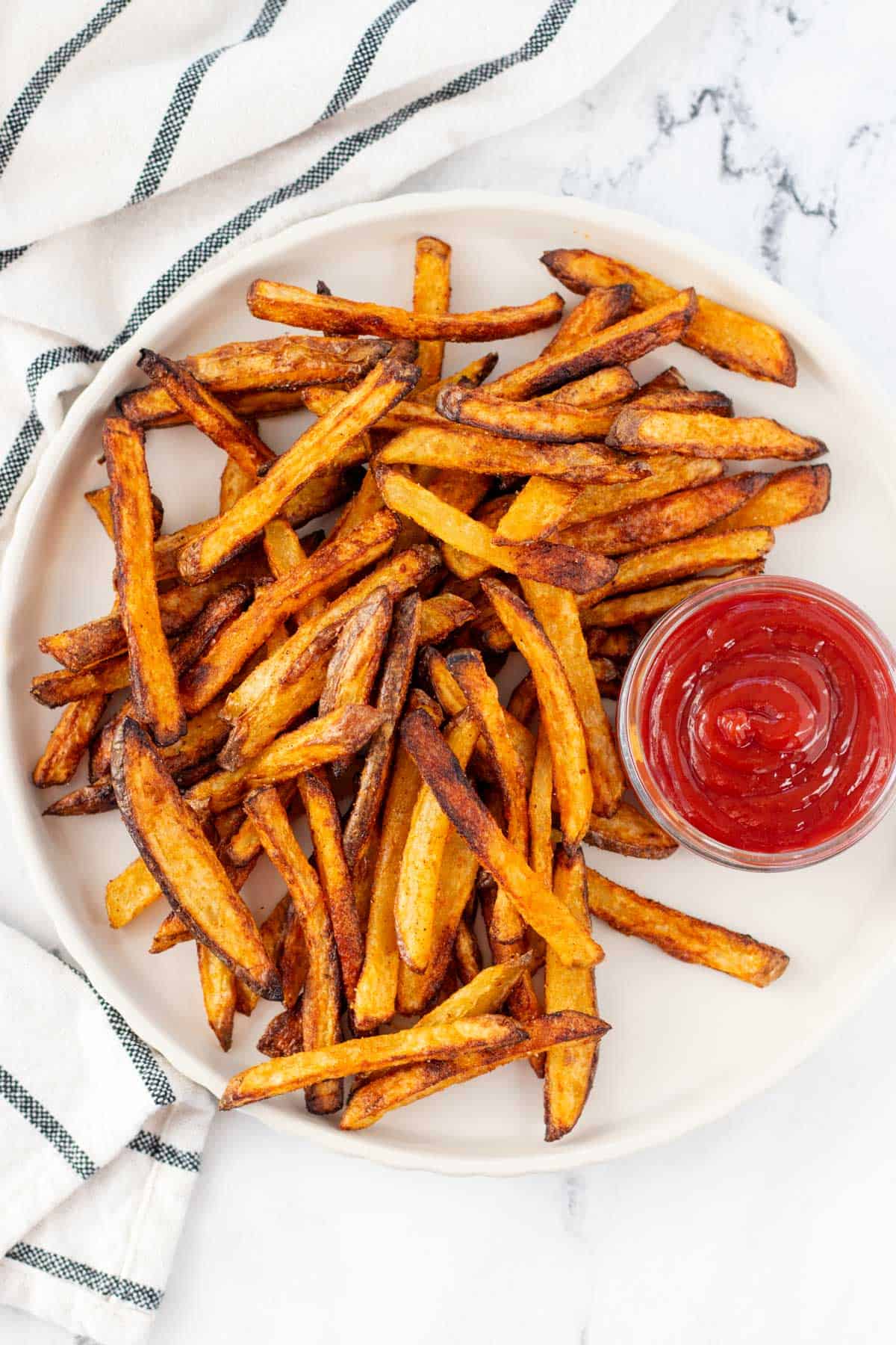 A plate of golden, crispy baked french fries with a side of ketchup on a marble background.