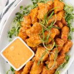 A white serving plate filled with bang bang chicken on a bed of arugula sprouts and a small dish of bang bang dipping sauce on the side.
