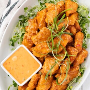 A white serving plate filled with bang bang chicken on a bed of arugula sprouts and a small dish of bang bang dipping sauce on the side.