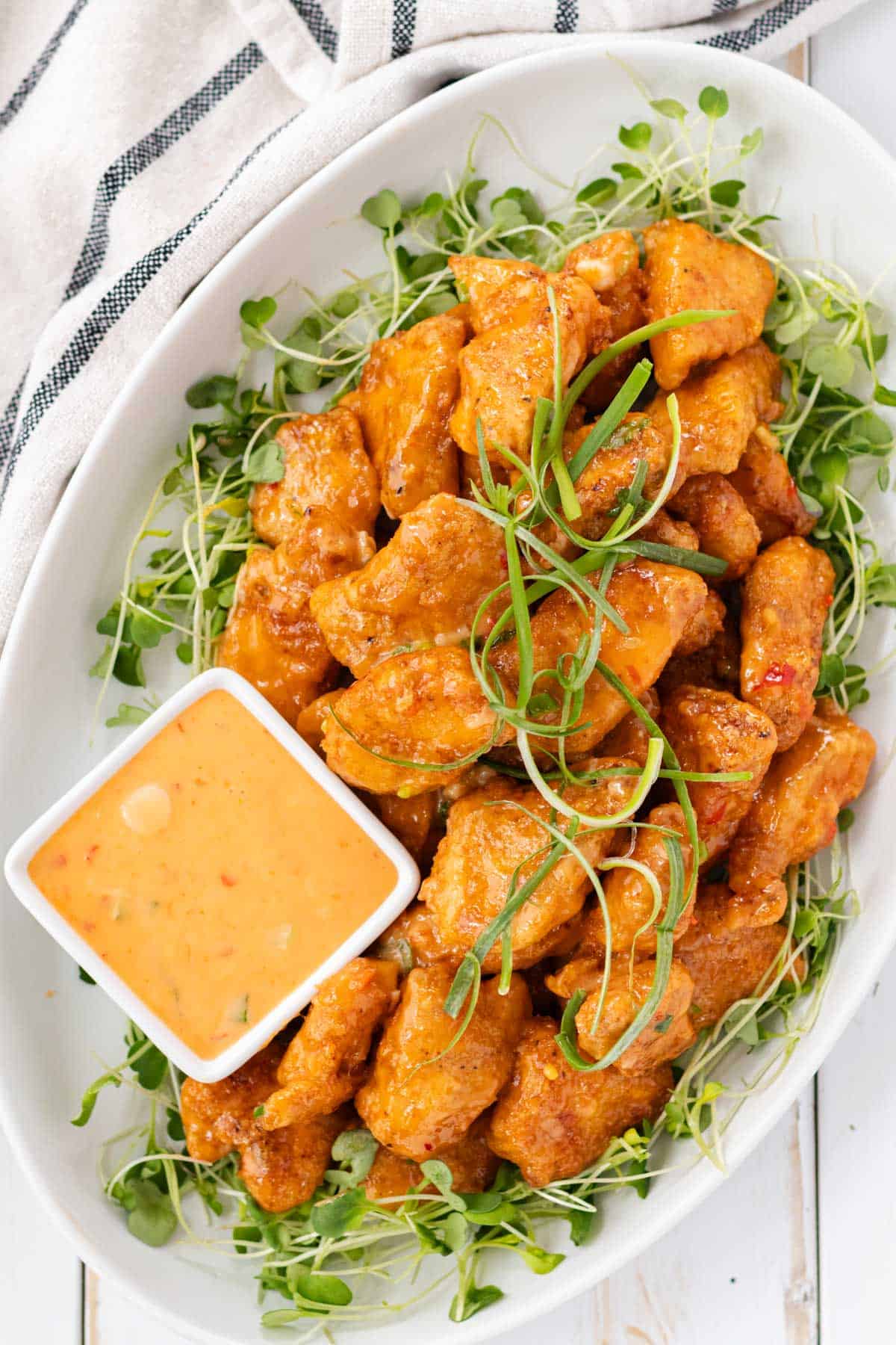 A white serving plate filled with bang bang chicken on a bed of arugula sprouts and a small dish of bang bang dipping sauce on the side.