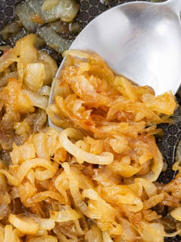 A silver spoon scooping golden brown caramelized onions from a pan.