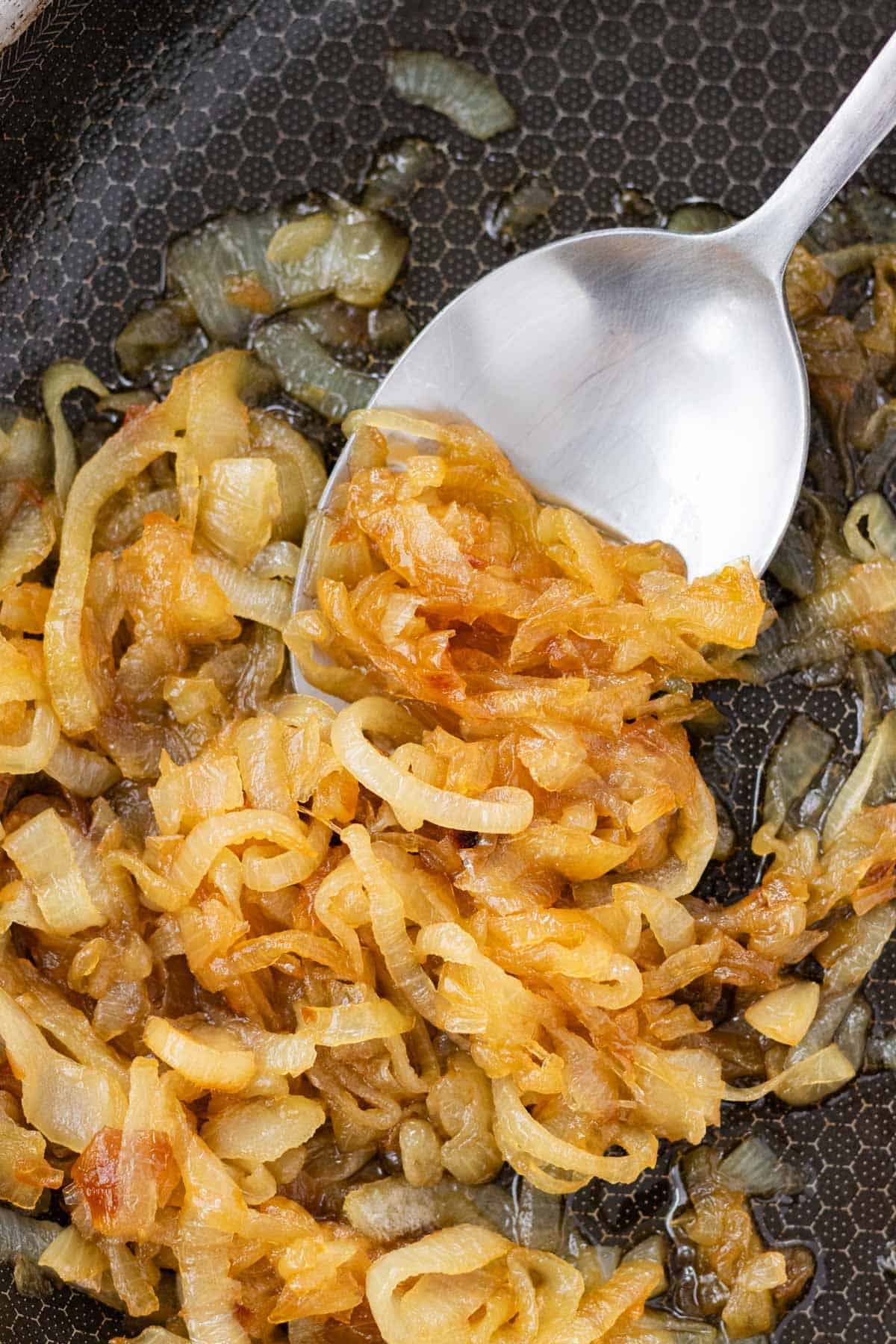 A silver spoon scooping golden brown caramelized onions from a pan.
