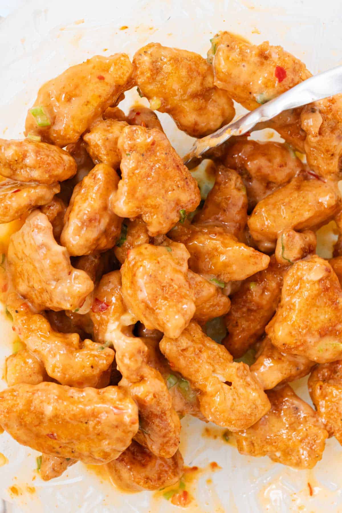 Tossing the chicken with bang bang sauce using a pair of tongs in a glass bowl.