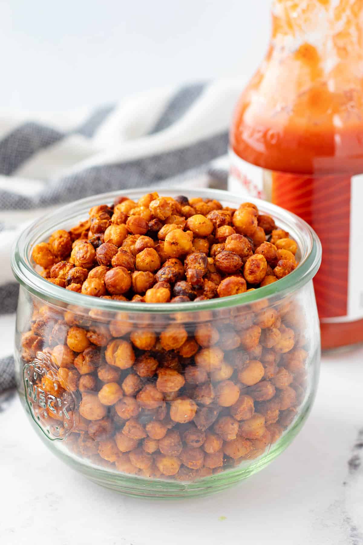 A jar full of crispy roasted buffalo chickpeas with a bottle of hot sauce in the background.