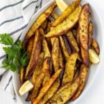 A white oval serving dish piled with greek lemon potato wedges with a sprig of parsley on the side and lemon wedges.