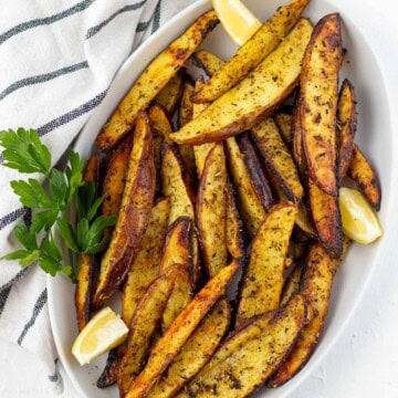 A white oval serving dish piled with greek lemon potato wedges with a sprig of parsley on the side and lemon wedges.