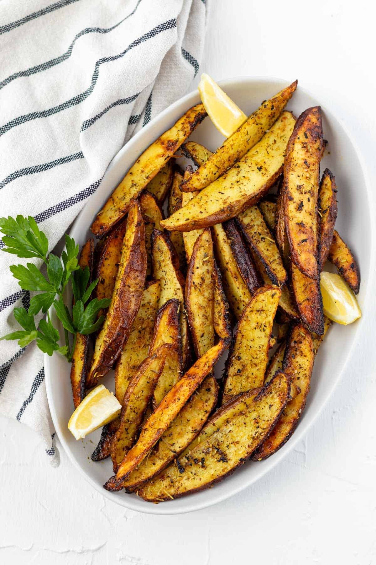A white oval serving dish piled with greek lemon potato wedges with a sprig of parsley on the side and lemon wedges.