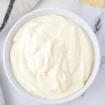 A white bowl full of avocado oil mayonnaise with a lemon and a dish towel on the side.