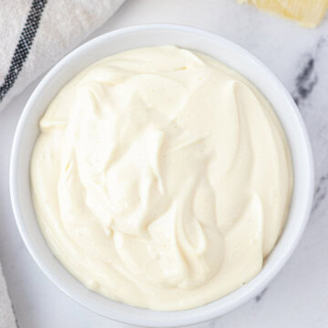 A white bowl full of avocado oil mayonnaise with a lemon and a dish towel on the side.