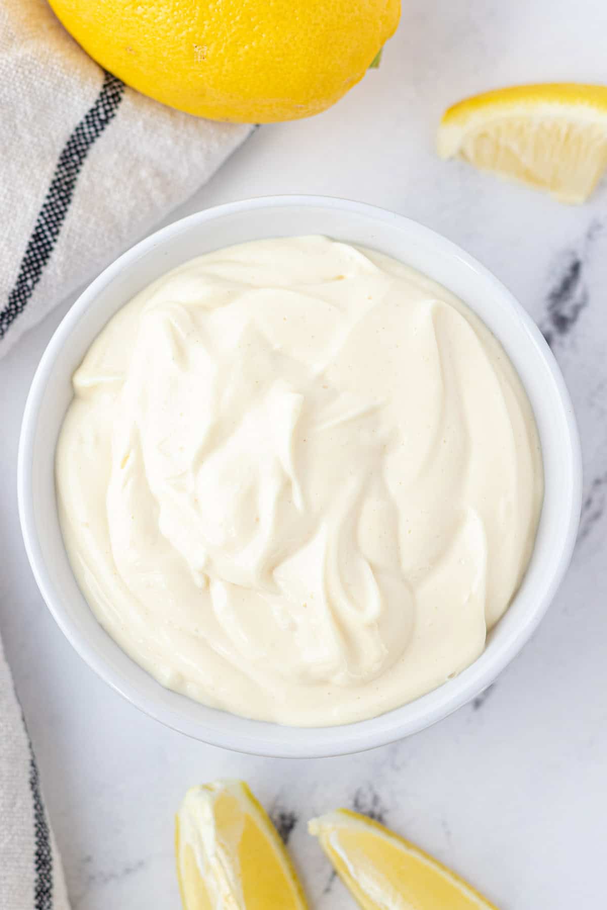 A white bowl full of avocado oil mayonnaise with a lemon and a dish towel on the side.