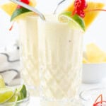 Two piña coladas with straws, a lime wedge, maraschino cherry, and pineapple wedge on the side.