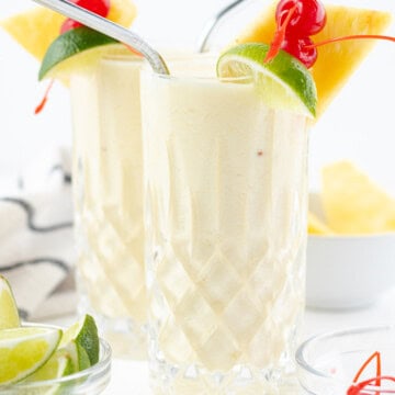 Two piña coladas with straws, a lime wedge, maraschino cherry, and pineapple wedge on the side.