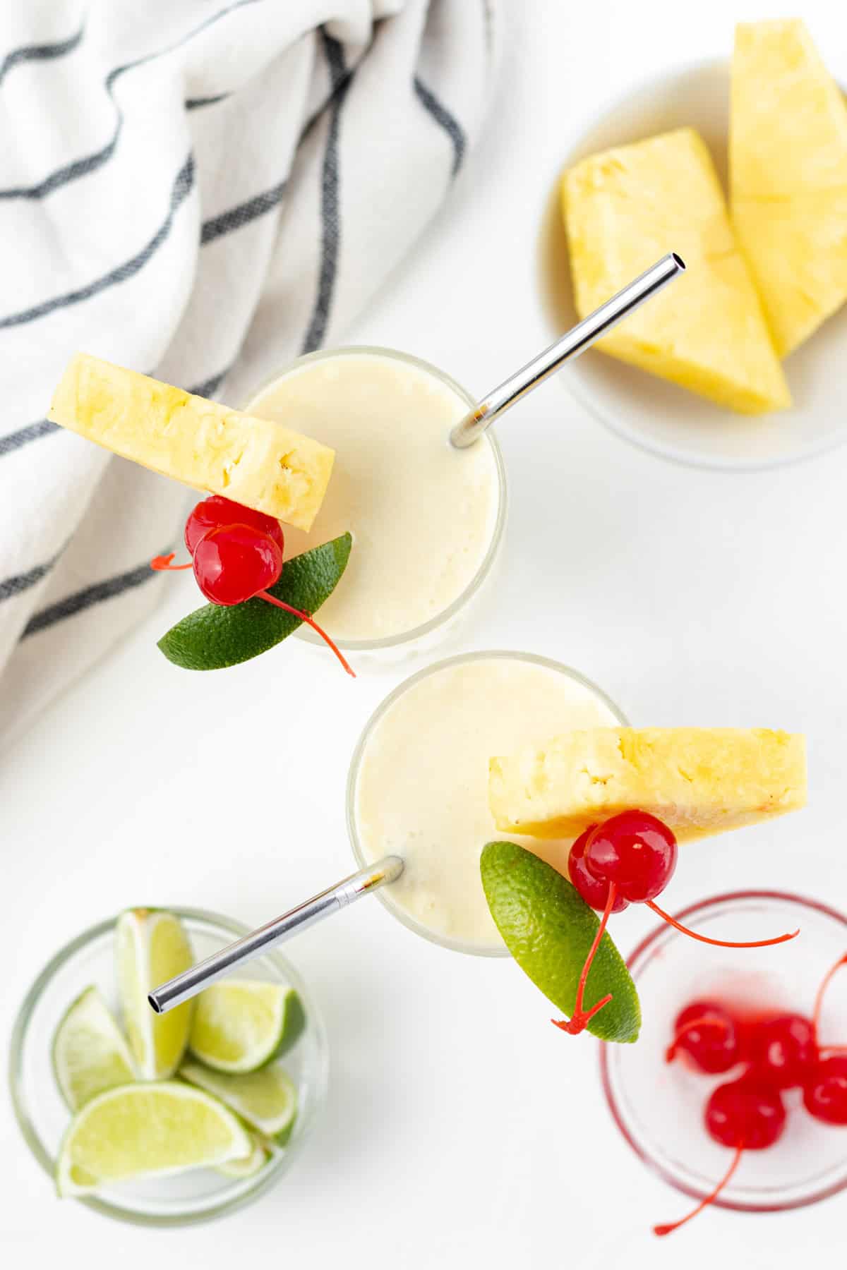 Two piña coladas in glasses with pineapple, lime and maraschino cherries on the side in dishes.