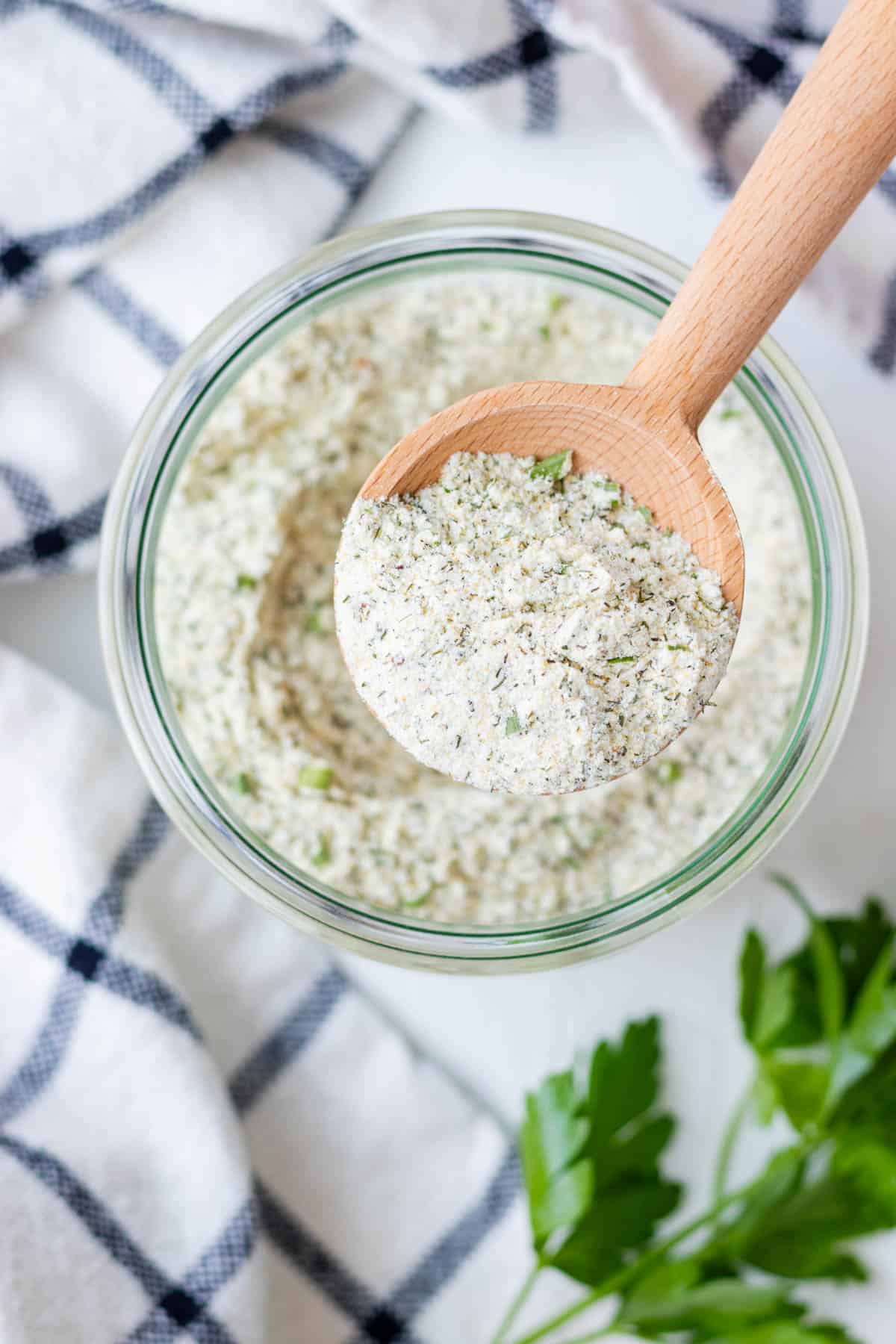 A round wooden spoon half full of ranch powder hovering over top of the jar.