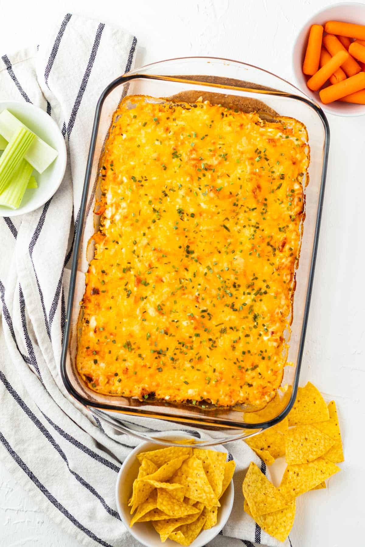 A pan of buffalo chicken dip after it has been baked with celery, carrots, and tortilla chips on the side.