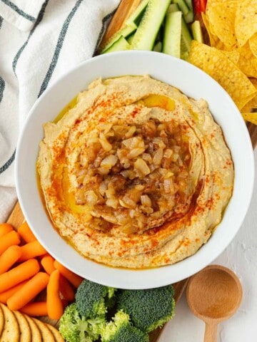 A white bowl filled with caramelized onion hummus with vegetables, chips, and crackers on the side.