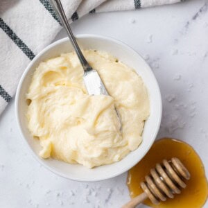 A white bowl filled with creamy whipped honey butter with a metal butter knife resting inside, placed next to a wooden honey dipper in a small puddle of honey.
