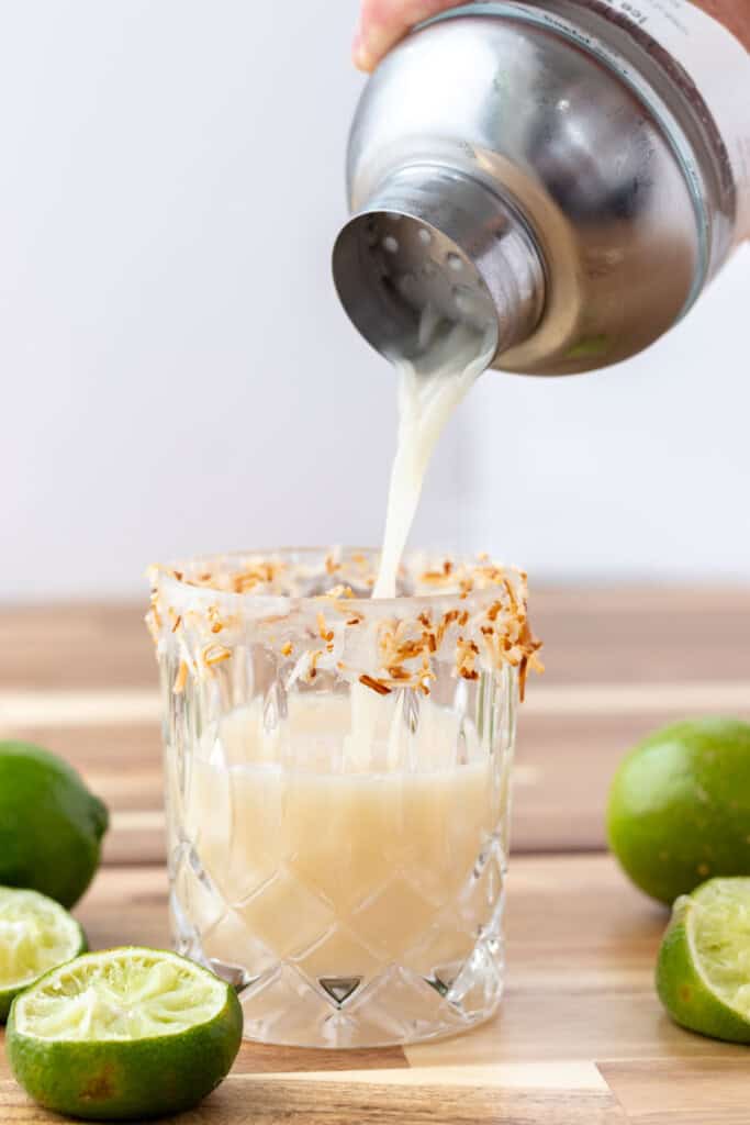 Pouring the shaken coconut margarita into a glass rimmed with toasted coconut.