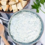 A blue serving bowl filled with spinach dip with a bowl of sourdough bread for dipping on the side.