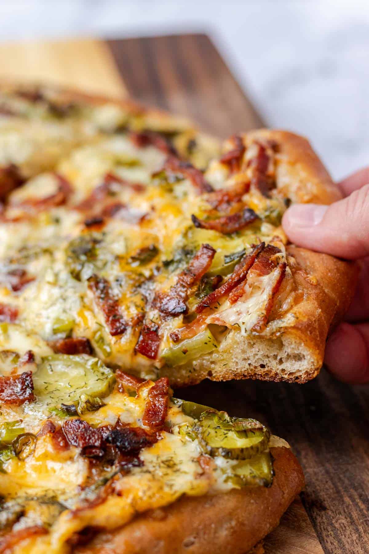 A hand grabbing a slice of dill pickle pizza off a cutting board.