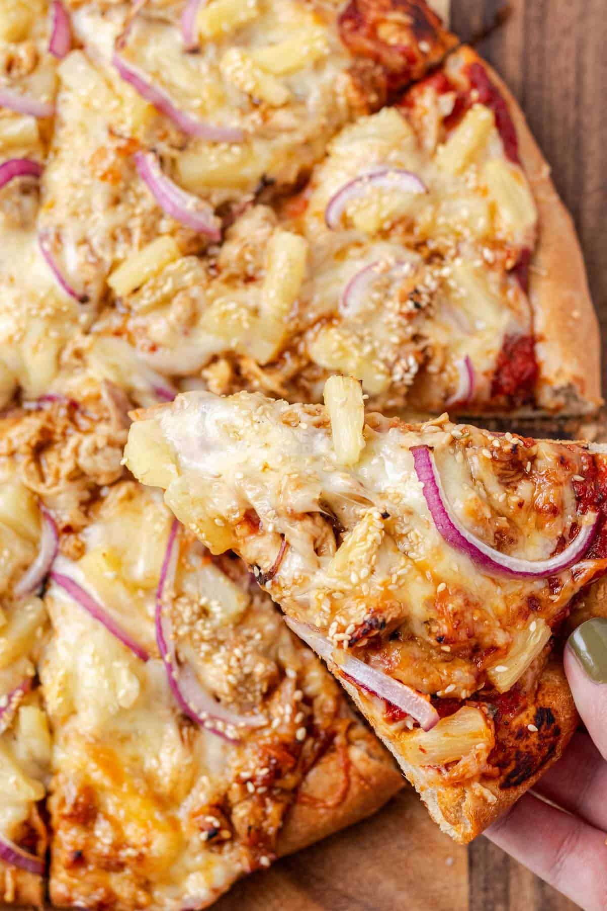 A hand pulling a piece of teriyaki chicken pizza from the whole pizza.